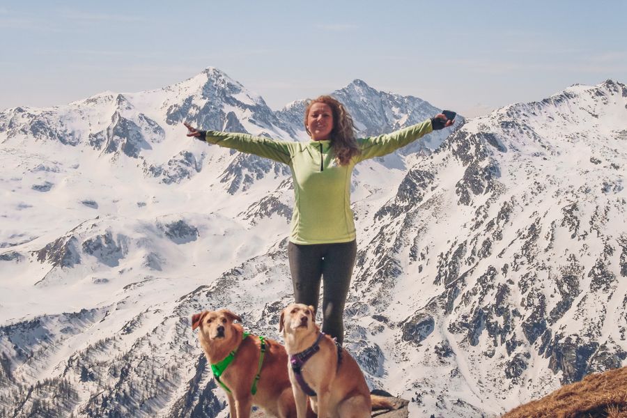 Randonneuse bras écartés devant des sommets enneigés, avec deux chiens en harnais à ses pieds