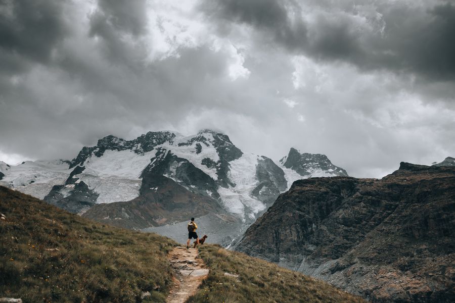 Randonneur et son chien sur un sentier de montagne face à des sommets enneigés